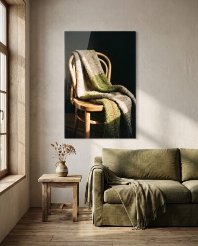 A folded wool blanket drapes over the curved back of a bentwood chair in low honeyed light, one rust thread threading through the muted green weave.