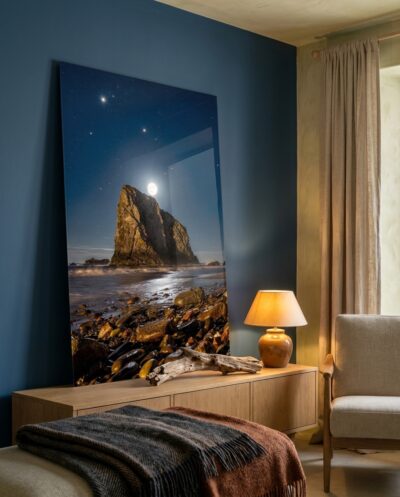 A towering coastal sea stack catches the radiant glow of a rising moon, illuminating wet beach stones beneath a brilliant starry sky.